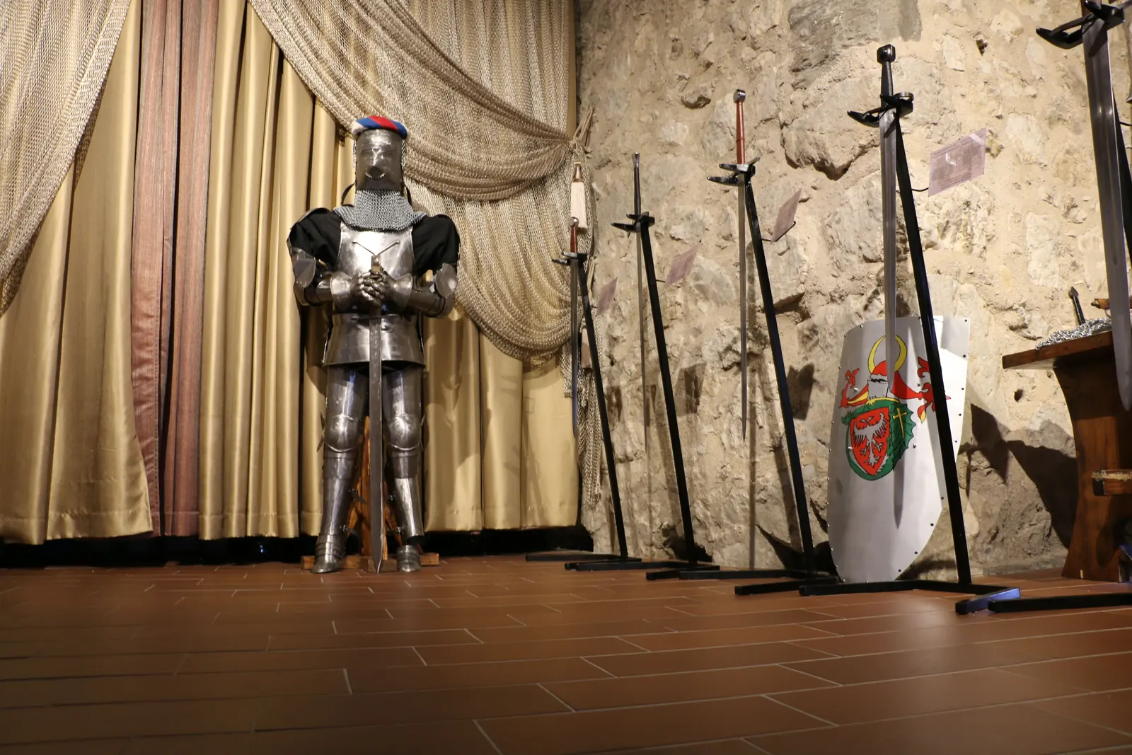 Un'armatura completa si erge accanto a spade in rastrelliera e uno scudo araldico contro una parete di pietra grezza, con una tenda dorata sullo sfondo