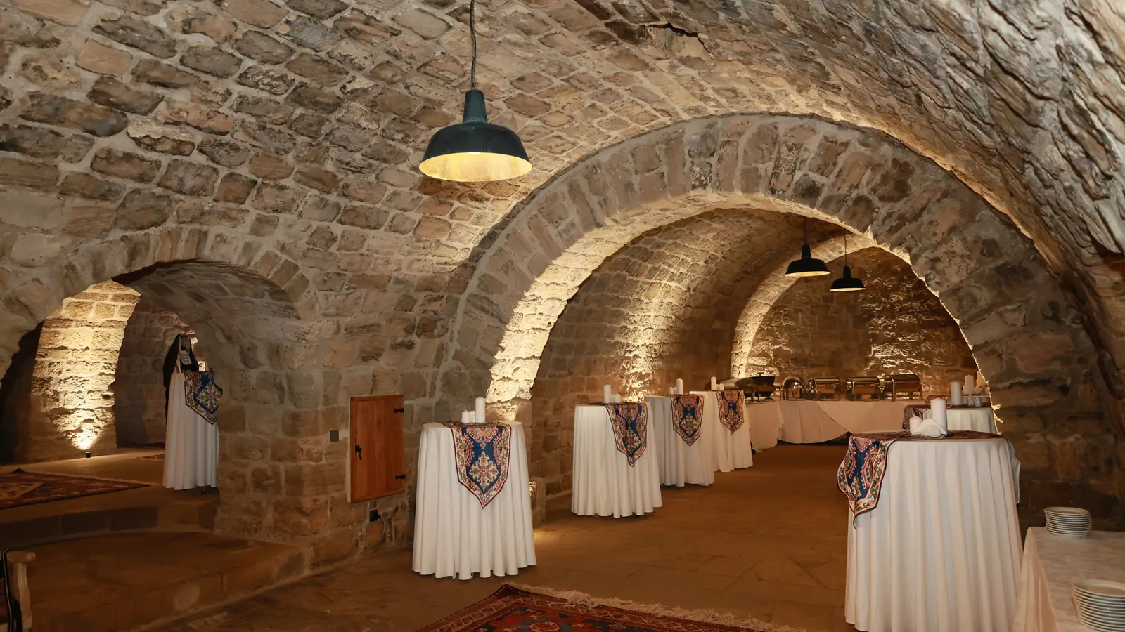 Una cantina in pietra a volta con soffitti ad arco, calde luci da parete e tavoli rotondi apparecchiati con tovaglie bianche per un banchetto