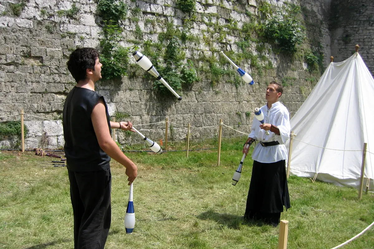 Due giocolieri in abiti di stile medievale lanciano clave davanti a un muro di pietra e a una tenda bianca a una fiera
