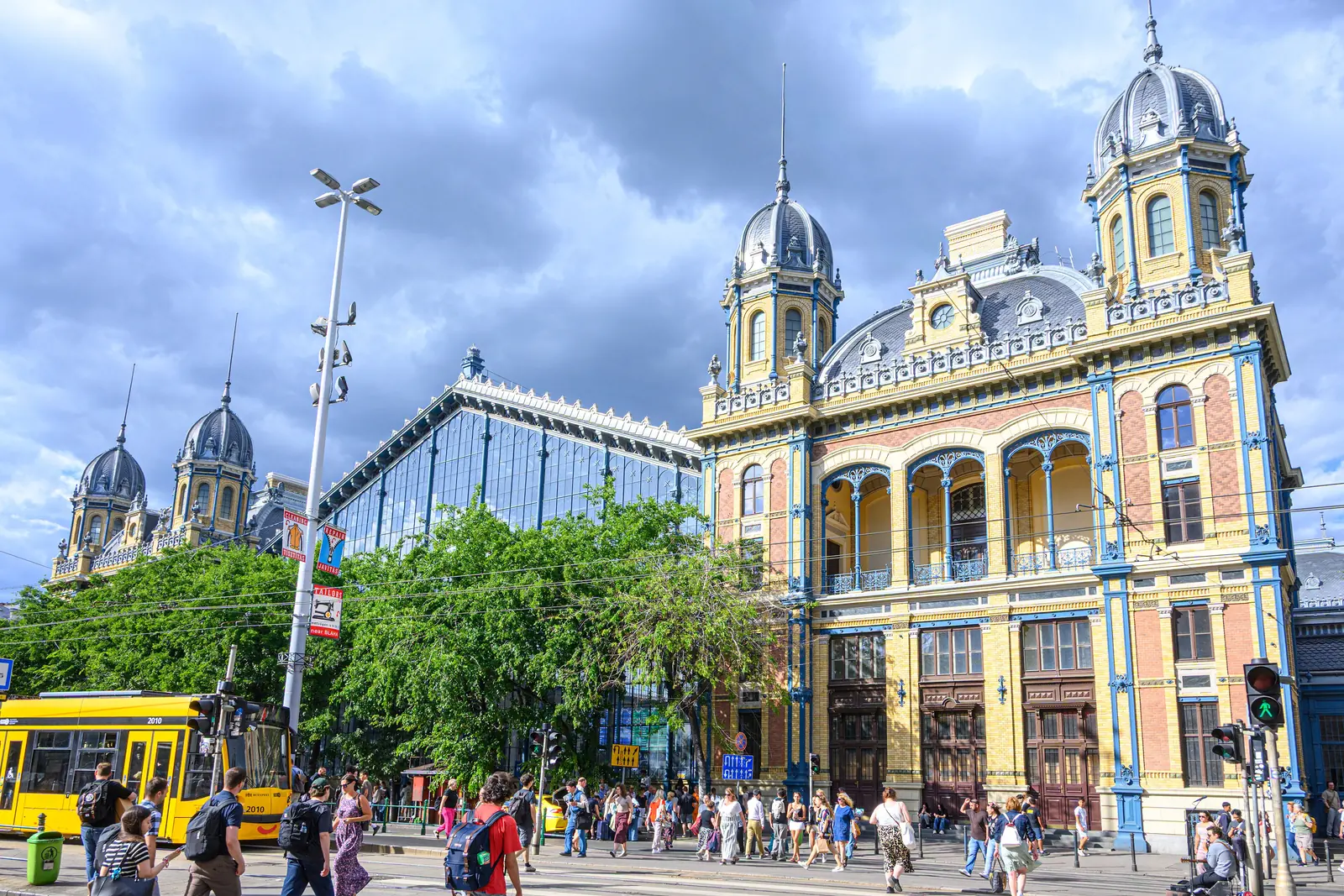 La facciata ornata gialla e blu della stazione Nyugati di Budapest con la sua sala in ferro e vetro, un tram giallo e la folla sul marciapiede