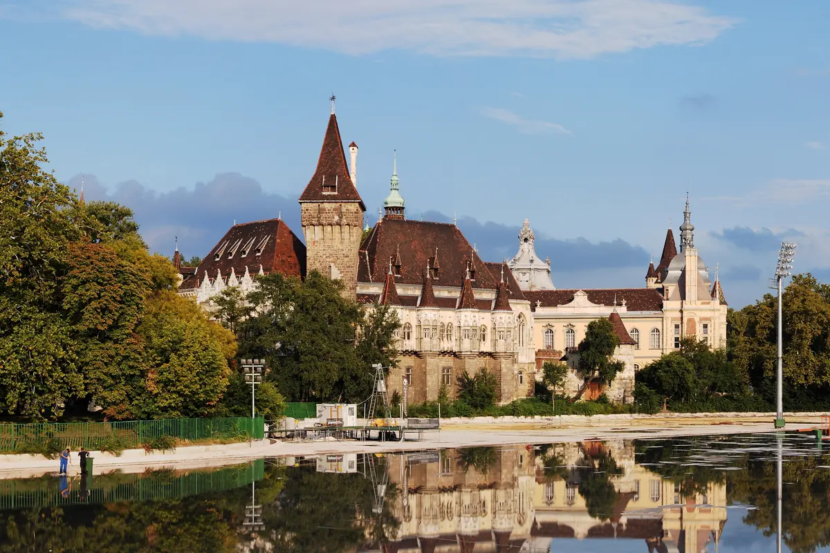 Il Castello di Vajdahunyad nel City Park di Budapest, un castello in mattoni e pietra con torrette riflesso nel lago sotto un cielo azzurro